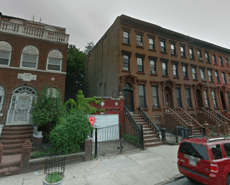 A brownstone building with stairs and a red car parked nearby.